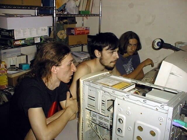 Ska from Milan, me in Bari, and Alk from Bologna, circa 2001 — three smanettoni who met on IRC and converged physically to do nerd things together. Open PC case, CRT monitors, ASUS boxes on the shelves. The natural habitat.