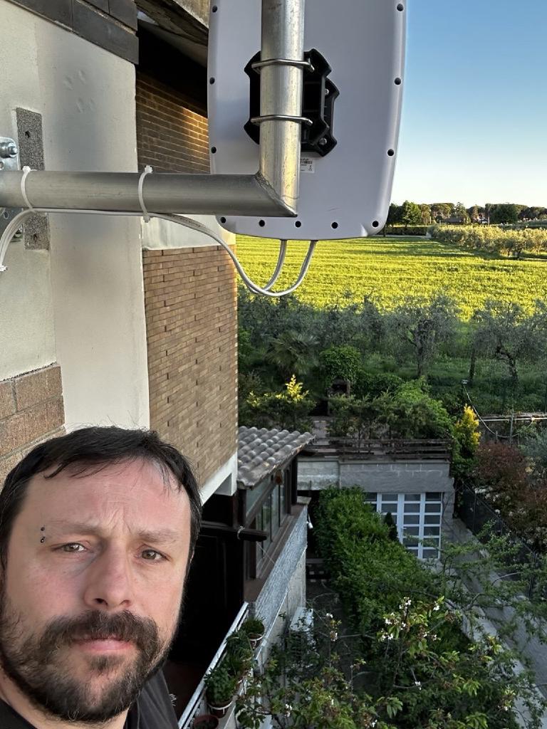 Selfie scattato dal basso — l'autore in basso a sinistra, sopra di lui l'antenna direzionale 5G montata sul muro dell'edificio, sullo sfondo campagna soleggiata e una vigna sotto un cielo limpido