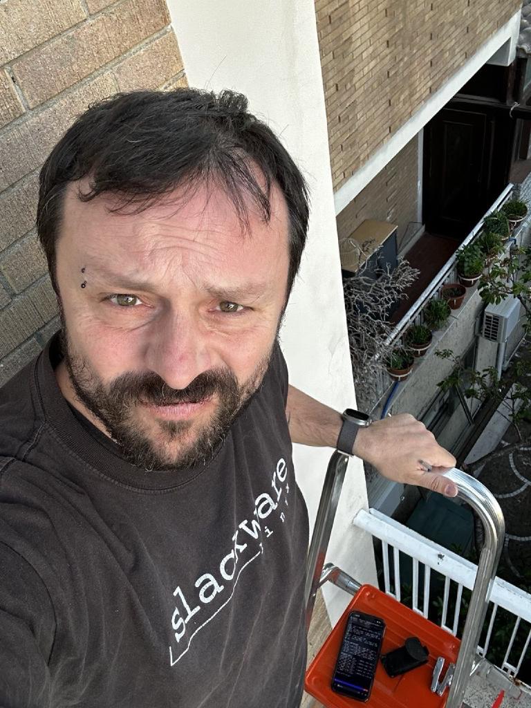 L'autore su una scala in piedi su un balcone, t-shirt Slackware, guarda dritto in camera. Un telefono che mostra l'output di 5g-monitor è appoggiato sul corrimano della scala accanto a lui; sullo sfondo un palazzo e qualche pianta in vaso