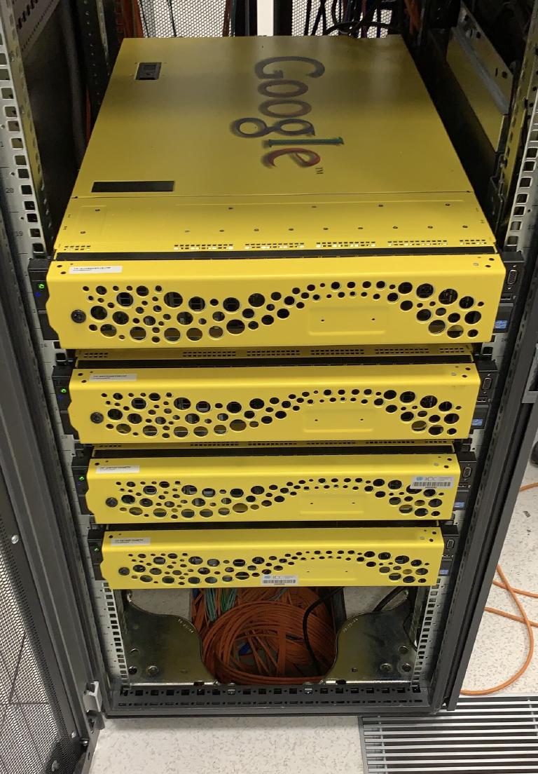 Four yellow Google Search Appliance servers stacked in a rack, each chassis embossed with the Google logo and perforated with the appliance's trademark pattern of round vents; orange fibre cabling visible behind them